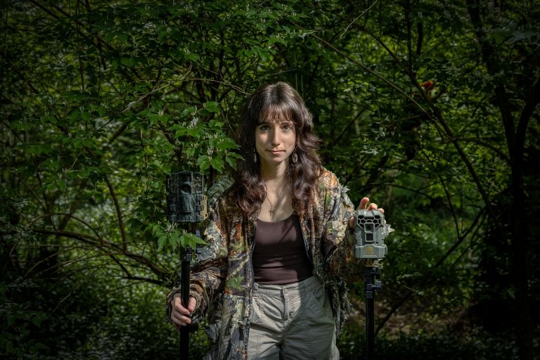 Arabella Fowler stands in a lush green forest, placing her hands on two cameras used to observe animals. She wears a camouflage jacket and light-colored trousers. The background features dense foliage and trees with a stuffed red panda in the background.