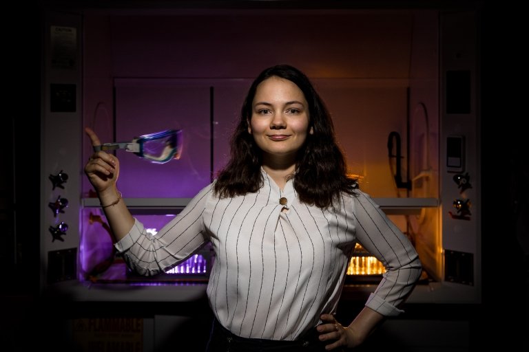 An stands confidently in a laboratory setting, twirling a pair of safety goggles in their right hand. The background features illuminated lab equipment with purple and orange lighting.