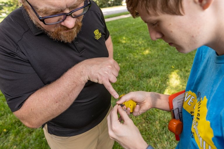 A student in a blue t-shirt holds a cicada shell as a man in a black polo shirt points to part of  the shell. 