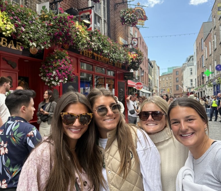 Four women smile for a photo in front of a building decorated with foliage.