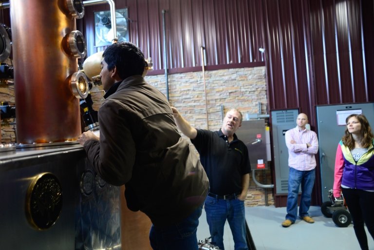 A young man peers into a glass porthole offering a view of the inside of a still at a bourbon distillery. 