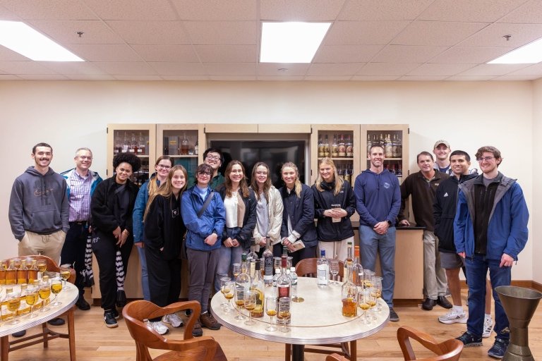 A large group poses for a photo standing behind tables filled with bourbon bottles for tasting. 
