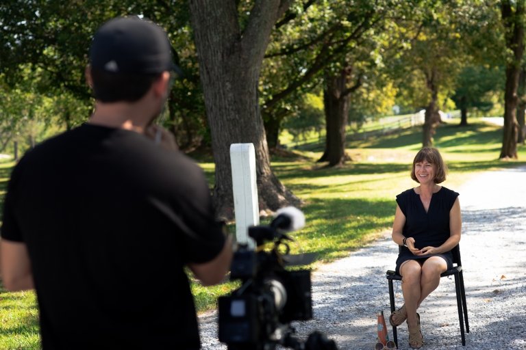 A man with a large camera on a tripod films a woman sitting outdoors as she is being interviewed. 