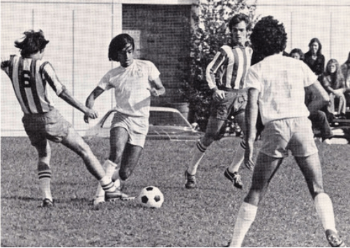 A black and white photo shows four men playing soccer, two wearing matching jerseys with vertical stripes and the other two wearing matching light-colored jerseys. 