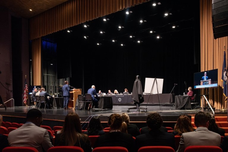 In the foreground audience members can be seen watching as court proceedings take place on a theater stage. 