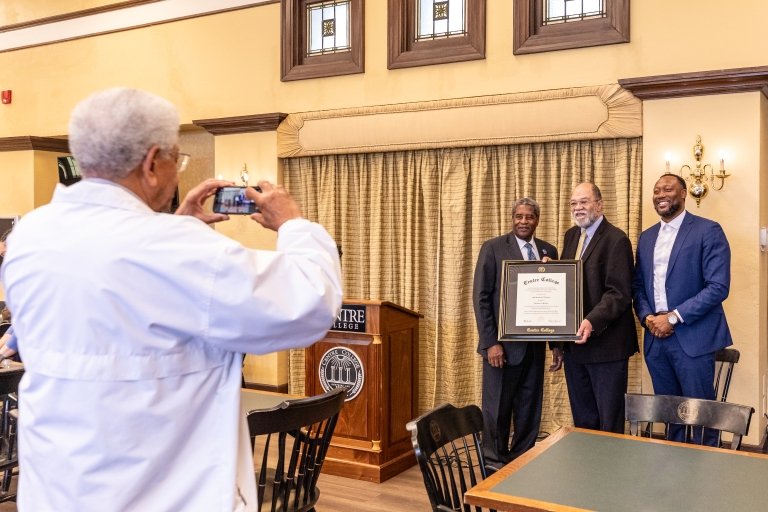 A man in a white coach takes a photo of three men posing together with a framed Centre College degree.