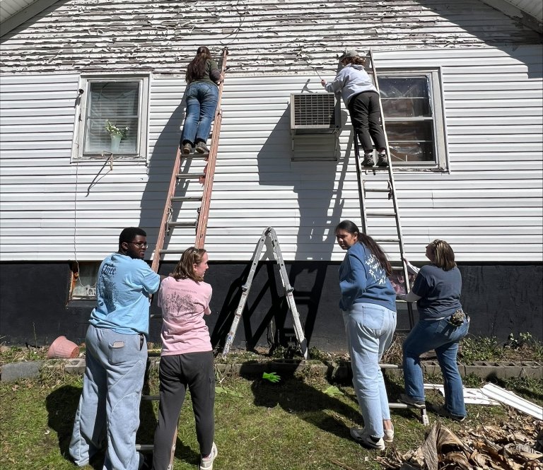 Two groups of students hold two step ladders as students at the top of each ladder prepare a house for new siding.