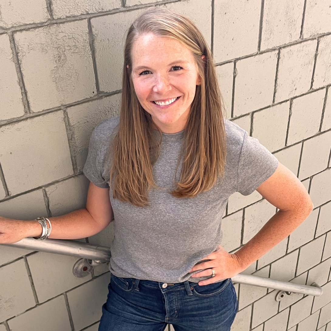 A woman wearing a grey t-shirt smiles for the camera while posing against a tile wall. 