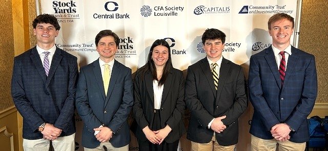 Two young men stand on either side of a young woman, all waring business suits and posing for a photo together. 