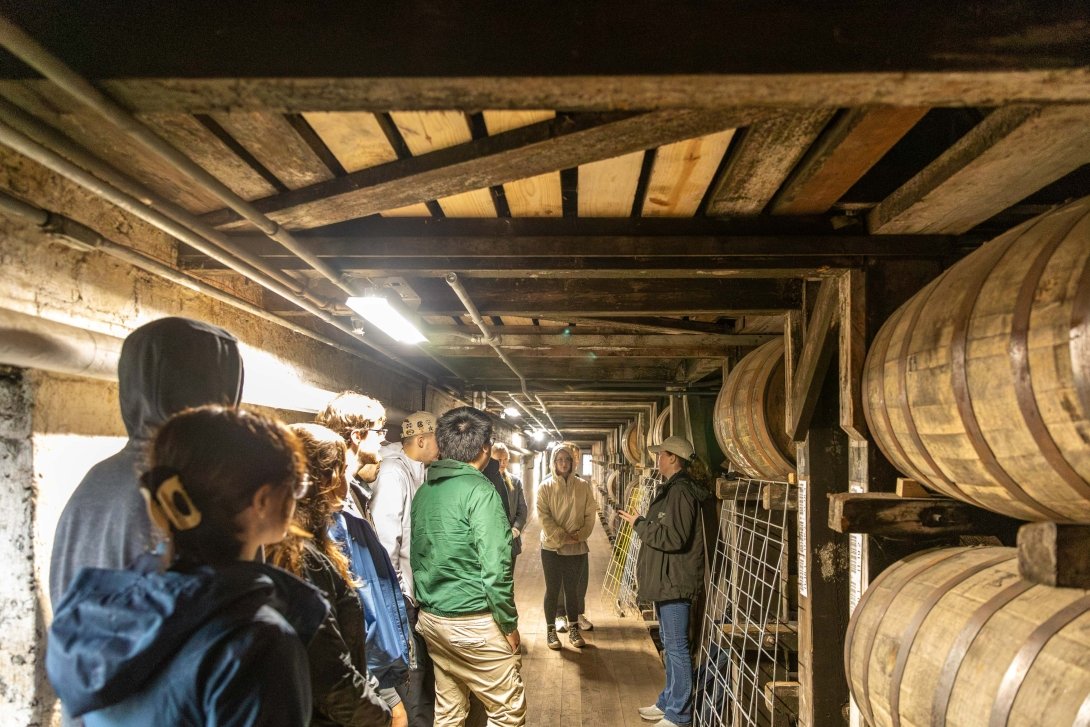 Students listen as to a man as they all stand surrounded by large wooden barrels in a bourbon aging warehouse, known as a rickhouse. 