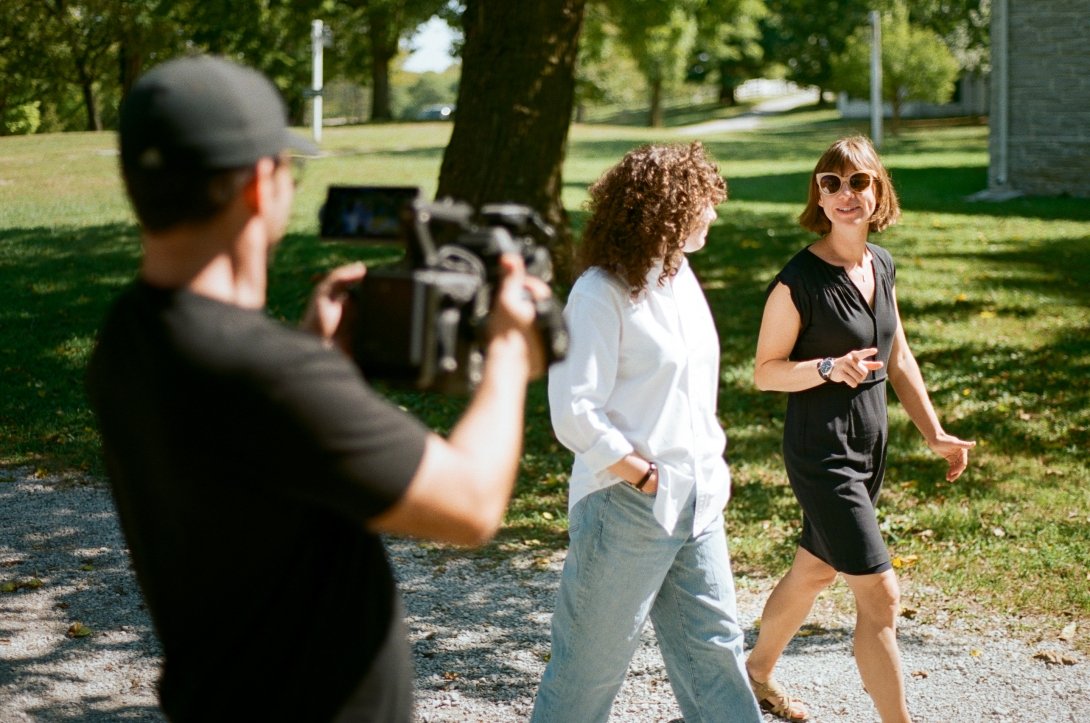 A man with a large camera videos two women walking and talking along an outdoor pathway. 