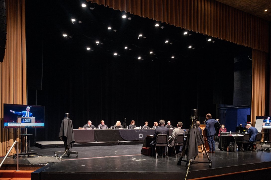 Kentucky Supreme Court Justices sit at long tables on a theatre stage as attorneys stand with their backs to the audience to argue their case. 